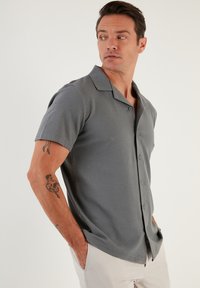 Camisa gris de manga corta y botones con una tela texturizada, cuello puntiagudo y dos botones frontales. Ajuste casual con bolsillos laterales.