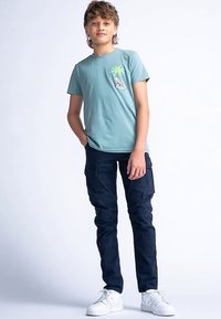 Světlomodré bavlněné tričko s grafikou palmy, doplněné tmavě modrými námořnickými cargo kalhotami. Outfit je dokončen bílými teniskami.