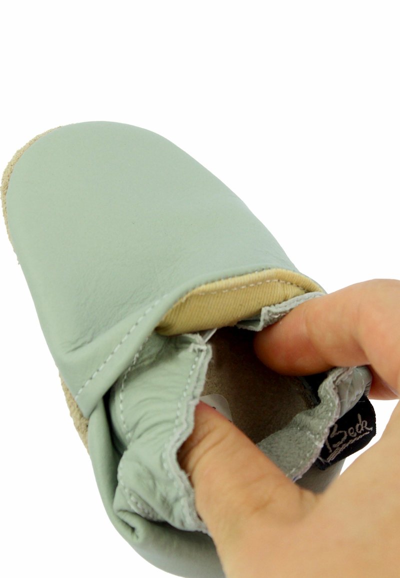 Main tenant une chaussure de bébé douce, vert clair, avec une ouverture élastique à la cheville sur fond blanc.
