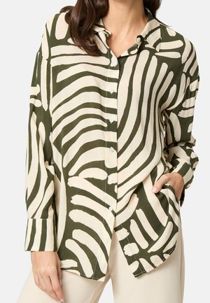 Frau trägt ein langärmliges Hemd mit auffälligen abstrakten dunkelgrünen und cremefarbenen Streifen, eine Hand in der Tasche, kombiniert mit einer hellen Hose.