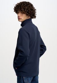 Tumesinane fleece'i tõmblukkjakk kõrge kraega, kahe küljetaskuga ja tekstuurse pinnaga. Mudel kannab seda siniste denimipükste peal.