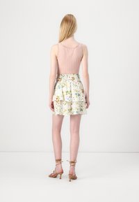 Jeune femme aux longs cheveux blonds portant un haut rose sans manches, une jupe à volants florale et des talons à brides beige, vue de dos sur fond blanc.