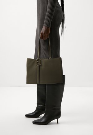 Oliven grøn læderhåndtaske med et struktureret design, flad form, guldfarvet hardware og dobbelte top-håndtag, holdt af en person.