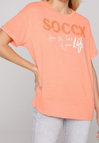 Camiseta de color durazno en tejido suave, con mangas cortas y un corte holgado. En la parte delantera tiene texto metálico en naranja y blanco con frases motivacionales.