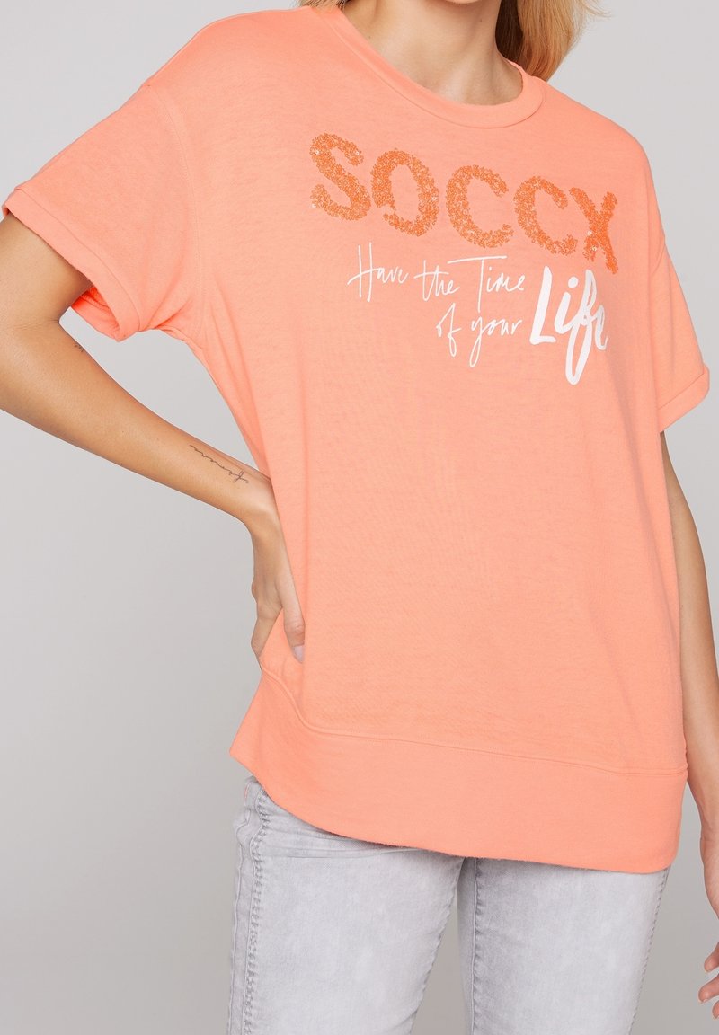 Camiseta de color durazno en tejido suave, con mangas cortas y un corte holgado. En la parte delantera tiene texto metálico en naranja y blanco con frases motivacionales.