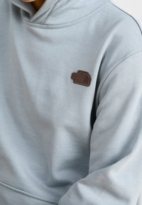 Světle modrá mikina s kapucí vyrobená z měkké látky, se strukturovanou hnědou nášivkou na hrudi a žebrovanými manžetami.