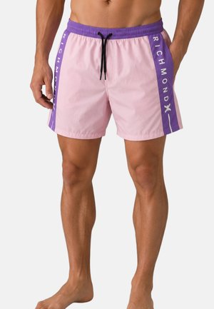 Uomo che indossa pantaloncini da bagno rosa con pannelli laterali viola e scritta "RICHMOND", in piedi a piedi nudi su sfondo bianco.
