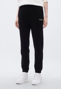 Μαύρο φόρμας με ελαστική μέση, στενά πόδια και κεντημένη λεπτομέρεια λογότυπου. Συνδυασμένο με λευκά αθλητικά παπούτσια. Μαλακή υφή υφάσματ ος.