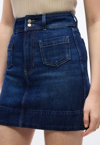 Mini-jupe en jean bleu foncé taille haute avec poches avant et fermeture double boutons, portée avec un haut côtelé beige.