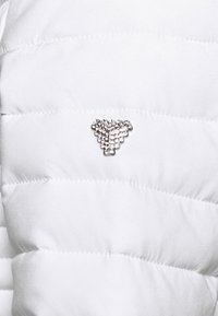 Logo triangulaire argenté composé de petits clous sur un fond en tissu matelassé blanc.