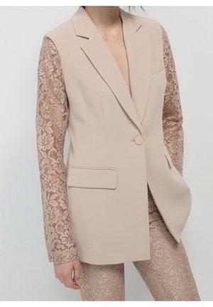Mujer con blazer beige a medida con mangas de encaje combinada con pantalones de encaje a juego, posando contra un fondo liso.