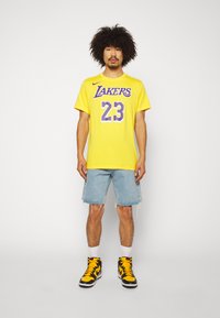 Gul bomulls t-skjorte med "Lakers" grafikk og nummer "23" i lilla. Brukt med lyse denimshorts og svart/gule joggesko.