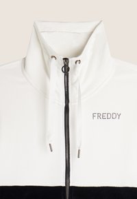 Veste zippée blanche avec panneau inférieur noir, dotée d'un col haut, de cordons de serrage et d'un logo en strass "FREDDY" sur la poitrine. Texture douce.
