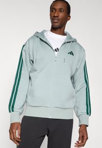 Φωτεινή γαλάζια ζακέτα με φερμουάρ, κατασκευασμένη από μαλακό ύφασμα. Διαθέτει σκούρες πράσινες ρίγες κατά μήκος των μανικιών και ένα μικρό λογότυπο στο στήθος. Απλός σχεδιασμός.