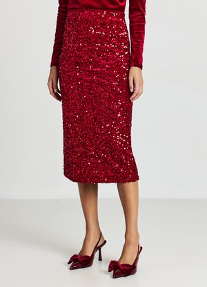 Gonna in paillettes rosse con silhouette aderente e lunghezza al ginocchio. Abbinata a décolleté in velluto rosso con dettagli a fiocco.
