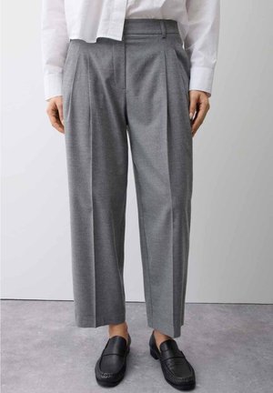 Pantalon plissé gris de longueur moyenne porté avec des mocassins noirs et une chemise blanche à manches longues sur un fond uni.