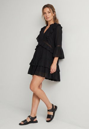 Femme debout contre un mur uni, vêtue d'une robe noire à volants avec un décolleté en V, des manches évasées et des sandales noires à boucle.