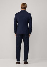 Granatowa marynarka o dopasowanym kroju, z pojedynczym rozporkiem i białymi detalami przy kołnierzu, zestawiona z ciemnymi dżinsami i brązowymi zamszowymi butami.