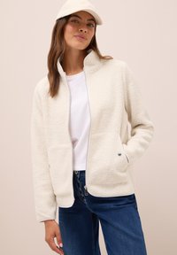 Femme portant une veste zippée en sherpa crème, un t-shirt blanc, un jean bleu et une casquette crème, posant sur un fond beige.