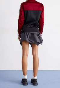Rote und schwarze Jacke mit schlankem Schnitt und gerippten Bündchen, kombiniert mit einem gestuften grauen Rock. Dazu werden schwarze Schuhe und weiße Socken getragen.