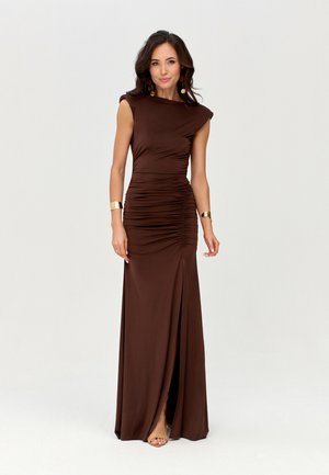 Vrouw die een mouwloze, gerimpelde, donkerbruine jurk tot op de vloer met een voorste split draagt, gouden armbanden, oorbellen en beige open-toe hakken.