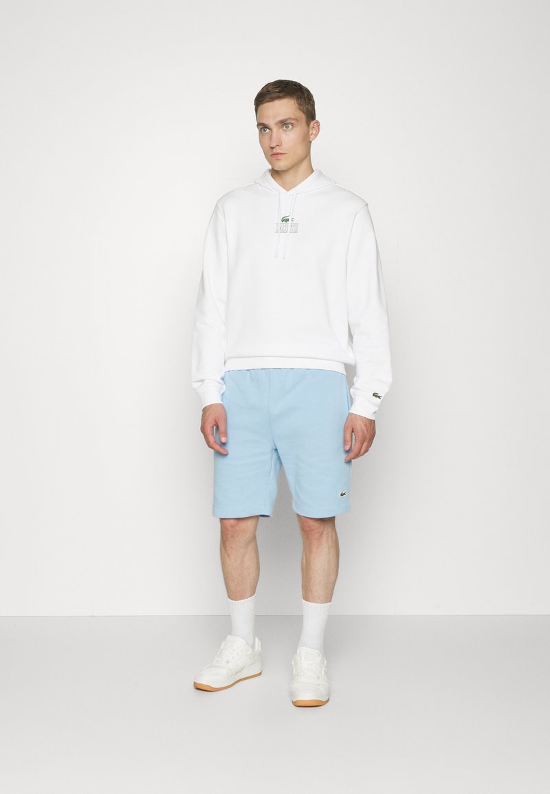 Hoodie blanc avec cordon de serrage, logo vert sur la poitrine. Shorts bleu clair, taille élastique. Baskets blanches avec semelle en gomme, chaussettes blanches.