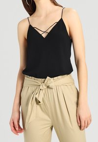Svart ärmlös topp med djup V-ringning och korsade axelband, kombinerad med beiga högmidjade byxor med knytband.