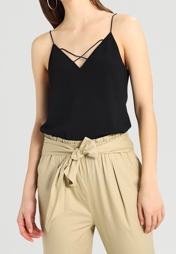 Svart ärmlös topp med djup V-ringning och korsade axelband, kombinerad med beiga högmidjade byxor med knytband.