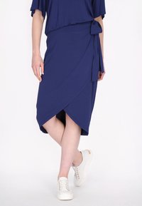 Navy blauwe wikkeljurk met een zijstrikdetail, gemaakt van zachte stof, met een diagonale zoom en gecombineerd met witte sneakers.