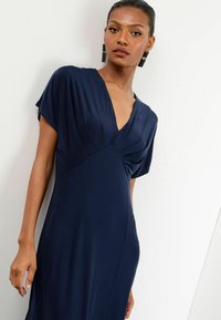 Navyblaues Kleid mit tiefem V-Ausschnitt, kurzen Ärmeln und gerafftem Detail an der Taille. Glatte Textur, taillierte Silhouette, knielange Länge.