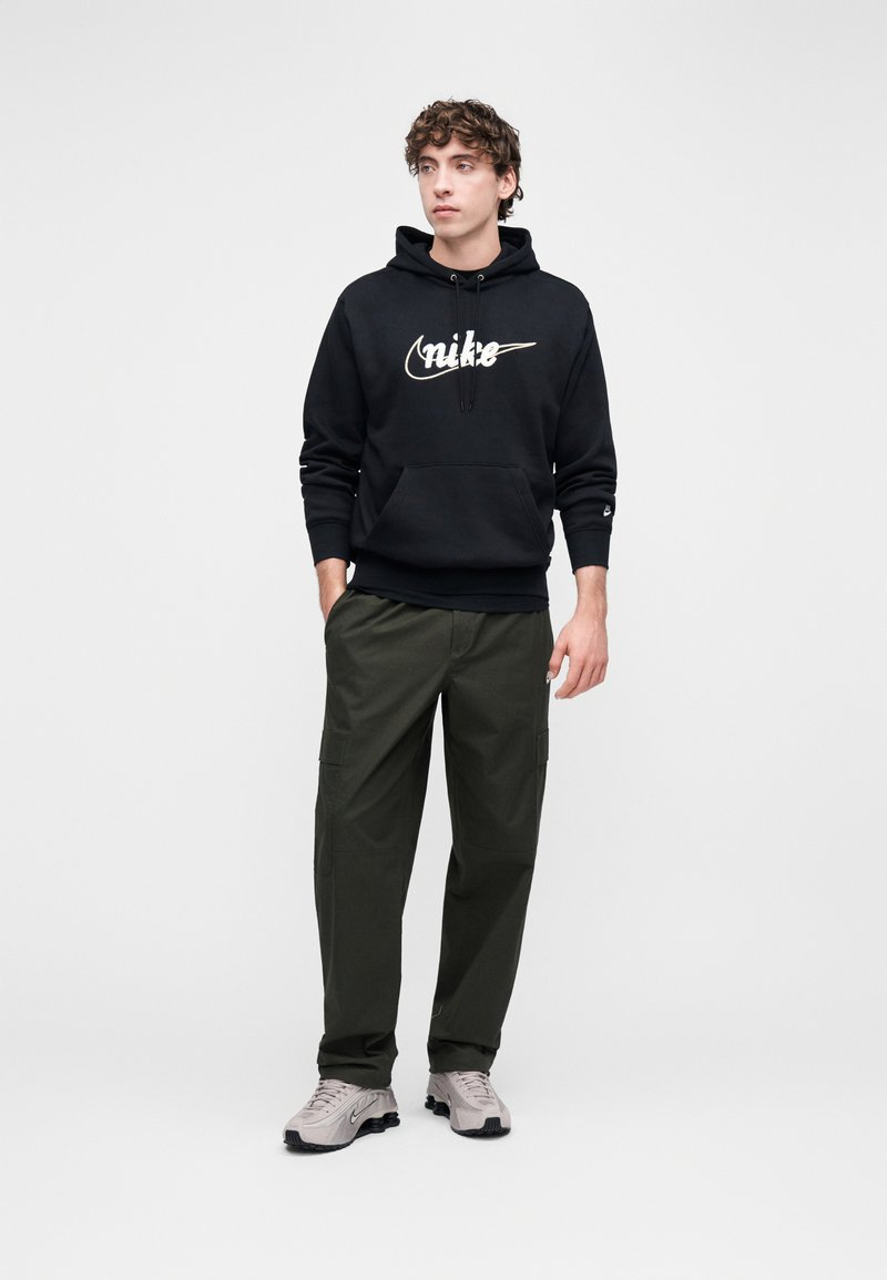Black hooded sweatshirt with prominent white "Nike" logo, paired with olive green cargo pants and light gray athletic shoes.