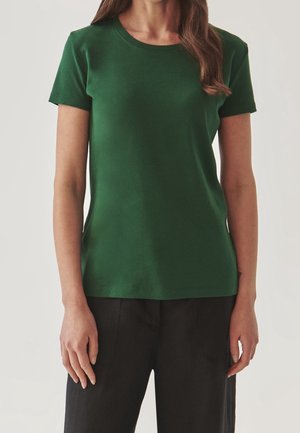 Kobieta ubrana w gładką, ciemnozieloną koszulkę z krótkim rękawem i okrągłym dekoltem oraz czarne spodnie, stojąca na neutralnym tle.