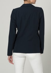 Kvinna i en figurnära marinblå blazer och vita byxor, sedd bakifrån mot en enkel ljus bakgrund.