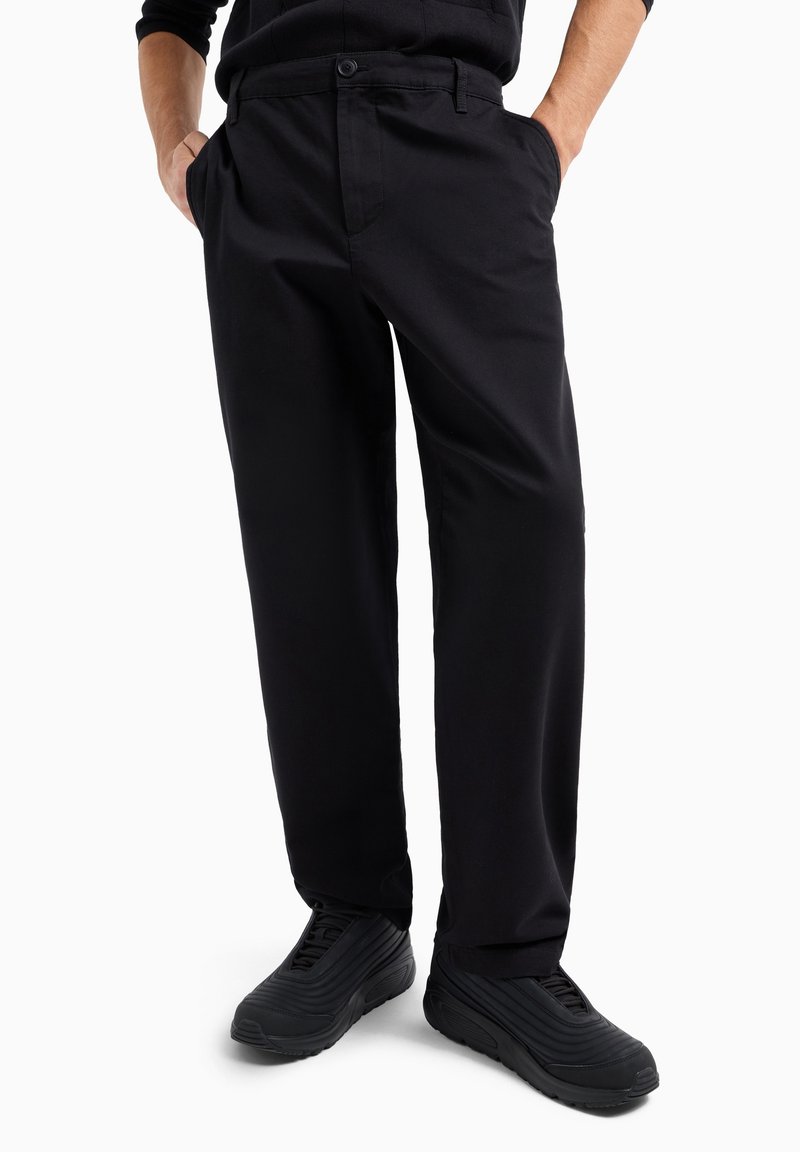 Pantaloni in cotone neri con una vestibilità comoda, dotati di tasche laterali e chiusura con bottone. Indossati con sneakers nere testurizzate.