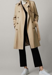 Beżowy trench z podwójnym zapięciem na guziki, dużymi klapami i paskiem. Noszony na czarnym golfie i czarnych spodniach, z białymi sneakami.