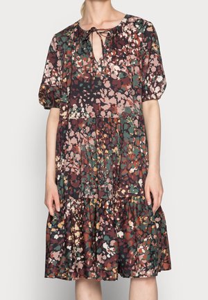 Kobieta ubrana w sukienkę do kolan z falbanami, bufiastymi rękawami i wielokolorowym abstrakcyjnym kwiatowym wzorem w stonowanych odcieniach ziemi.