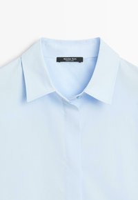 Hellblaues Hemd mit spitzem Kragen und schwarzem Markenetikett "Massimo Dutti" auf der Innenseite des Kragens.