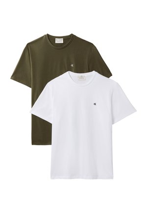 Mann steht mit den Händen in den Taschen, trägt ein weißes T-Shirt, ausgewaschene blaue Jeans, schwarzen Gürtel und schwarze Schuhe vor einem grauen Hintergrund.