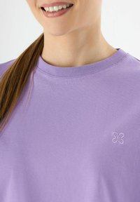 Paarse katoenen T-shirt met een ronde halslijn en korte mouwen. Bevat een kleine geborduurde bloemenaccent aan de voorkant. Glad textuur.