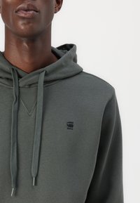 Hoodie vert foncé avec cordon de serrage, design en V, et un petit logo noir sur la poitrine. Texture lisse, coupe décontractée.