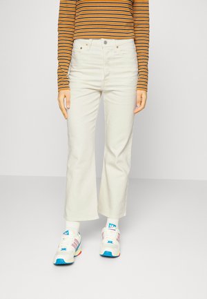 Personne portant un pantalon blanc taille haute à jambe droite, un haut à manches longues rayé orange et noir, des chaussettes blanches et des baskets blanches avec des accents bleus.