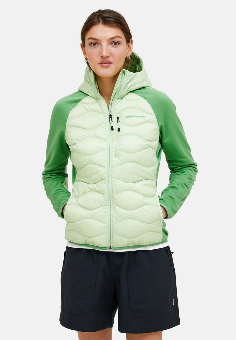 Giacca isolata verde chiaro con design trapuntato e cappuccio. Presenta maniche in un verde scuro contrastante e tasche con zip. I pantaloni abbinate sono scuri.