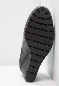 Zapato negro de cuero con una suela de goma texturizada que presenta surcos profundos y la inscripción "Calvin Klein Jeans" grabada. Colocado sobre una superficie plana.