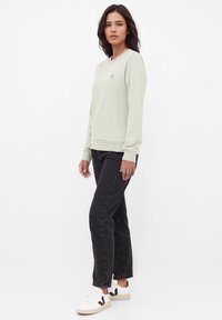 Hellgrüner Sweatshirt mit langen Ärmeln, Rundhalsausschnitt und gerippten Bündchen. Kombiniert mit schwarzen Jeans aus Denim und weißen Sneakers mit schwarzen Akzenten.