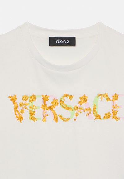 Hvit bomull t-skjorte med "VERSACE" logo dekorert med blomsterdesign i gule, grønne og rosa nyanser. Rund hals, korte ermer.