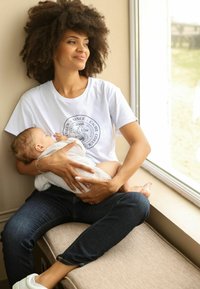 Witte T-shirt met een cirkelvormige afbeelding, blauwe jeans, met een in doeken gewikkelde baby in de armen, zittend bij een zonnig raam, zachte texturen en warme verlichting.