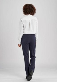 Witte langwerpige overhemd met plooidetail aan de achterkant, gecombineerd met marineblauwe broek met elastische taille en zwarte schoenen. De achtergrond is een effen lichtgrijze kleur.