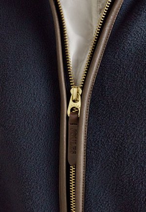 Nahaufnahme einer marineblauen Fleecejacke mit einem goldfarbenen Metallreißverschluss und braunen Lederbesätzen sowie einem Reißverschlussanhänger mit der Aufschrift „Joules.“