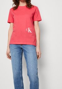 Korálová bavlněná trička s krátkým rukávem a kulatým výstřihem. Na dolním levém rohu je bílá logo CK. V kombinaci s modrými džínami.