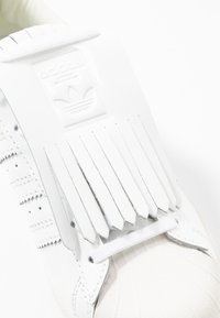 Vit sneaker med en framträdande fransad klaff, texturerat läder och design med tre ränder. Skosnören är vita och sulan är gjord av gummi.